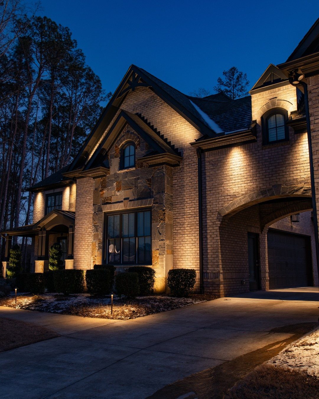 A large brick house illuminated by warm outdoor spotlights against a dark blue twilight sky.