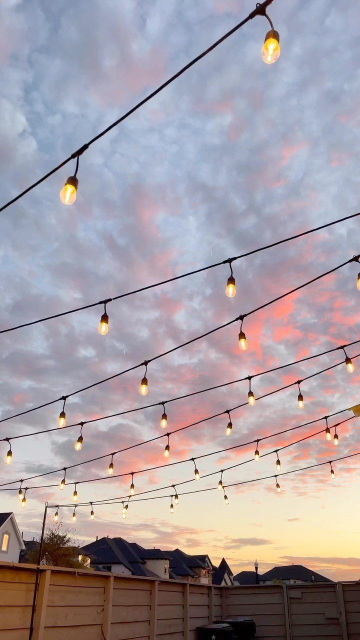 String lights with glowing bulbs hang across a backyard under a pink sunset sky.