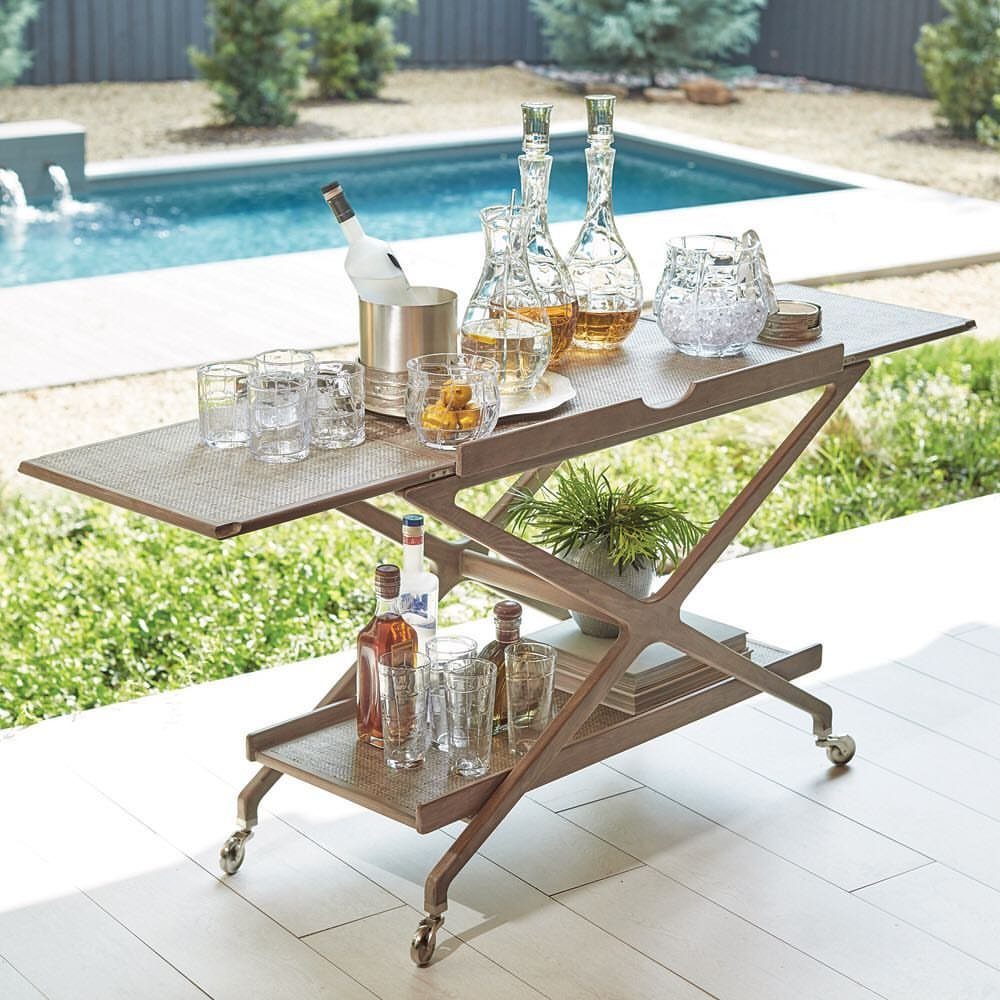 A metal two-tiered bar cart stocked with glassware and bottles on a white patio.