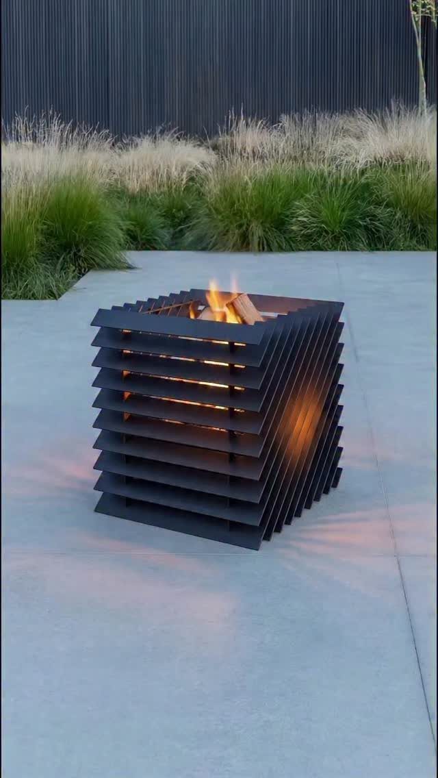 A modern black metal slatted fire pit burning on a smooth concrete patio.