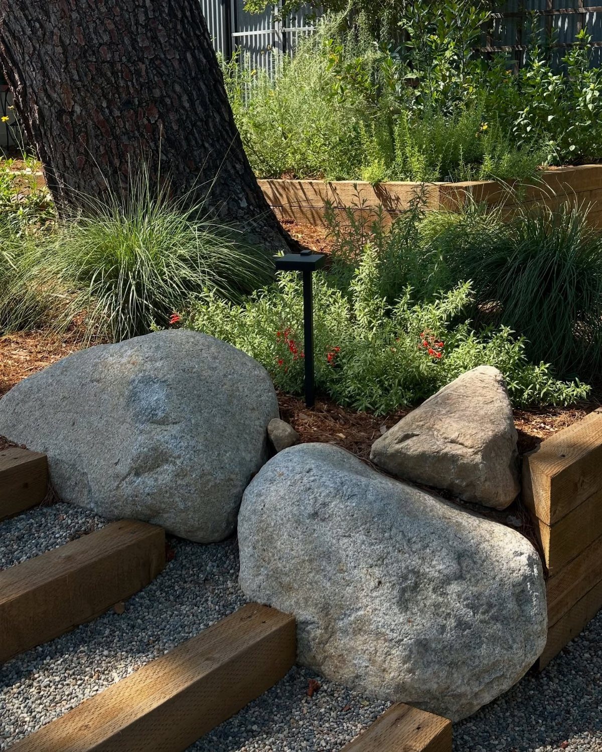 Large granite boulders serve as focal points in a landscaped garden bed with wood edging.