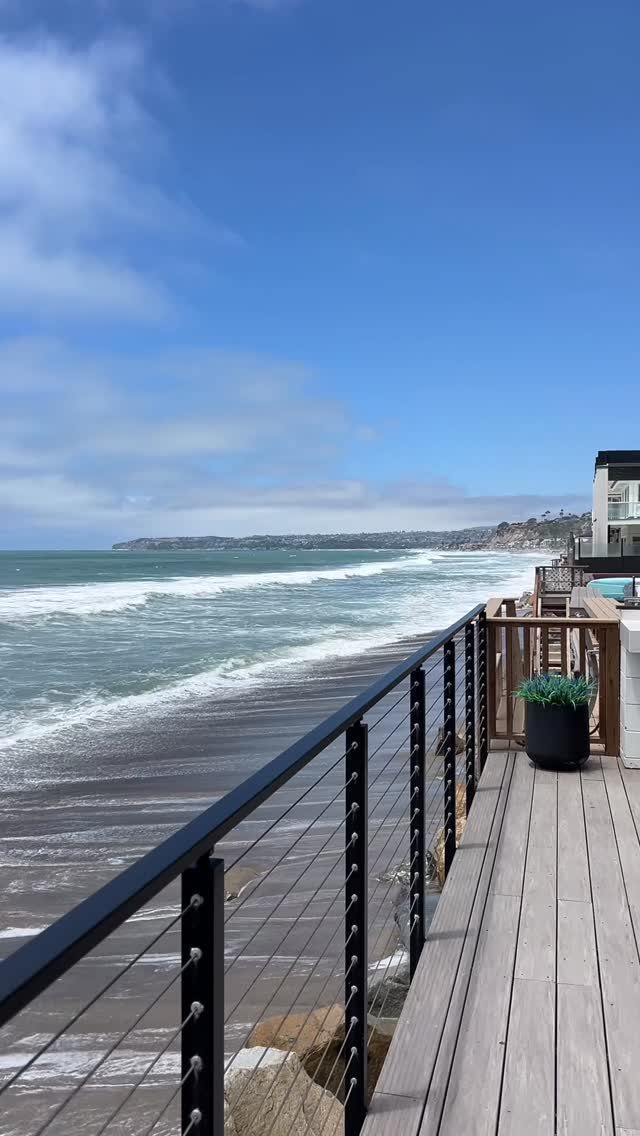 17 Beach House Deck Ideas for Oceanfront Living 15 A wooden deck with black cable railings overlooking a sandy beach and ocean waves.