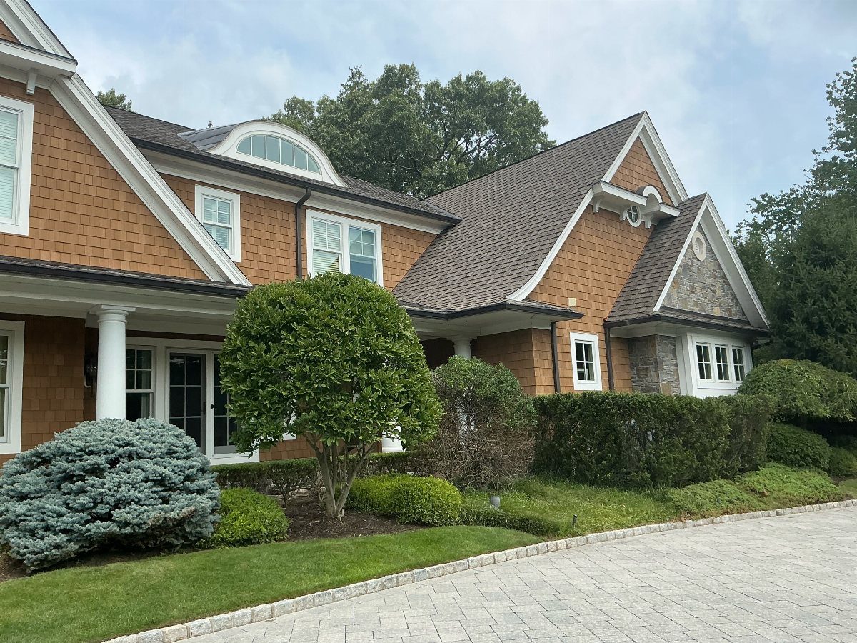 A large house with brown cedar shake siding and white trim surrounded by green bushes.