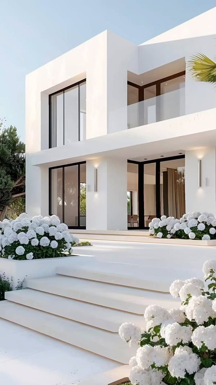 12 Trending House Colors for Exterior Looks in 2026 6 Modern white two-story house with large glass windows and white hydrangea flowers in the foreground.