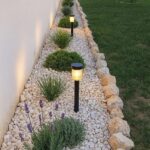 A narrow garden bed with white pebbles, green shrubs, and solar lights edged by natural stones.