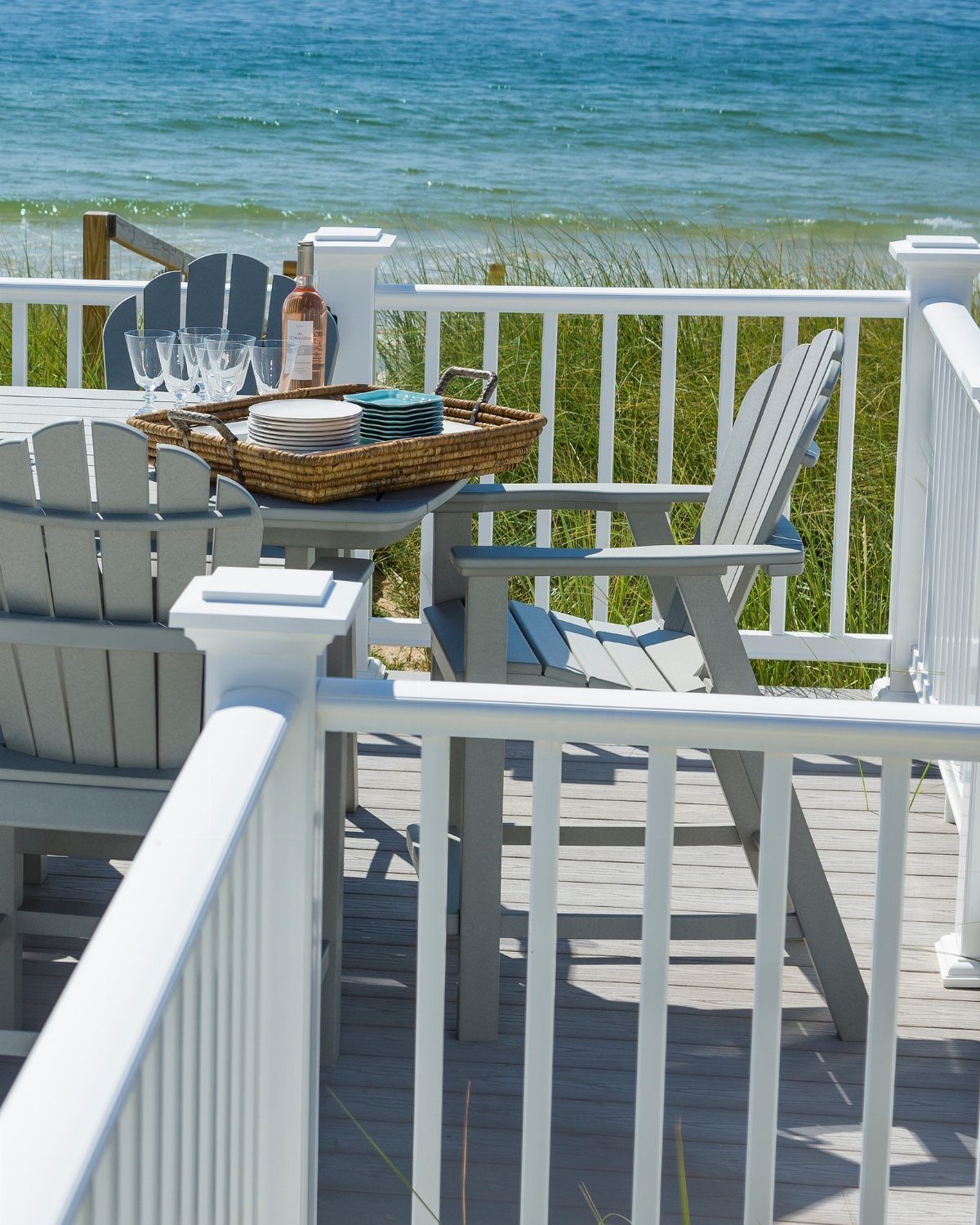 17 Beach House Deck Ideas for Oceanfront Living 7 Grey Adirondack chairs and a dining table on a white railed deck overlooking the ocean.