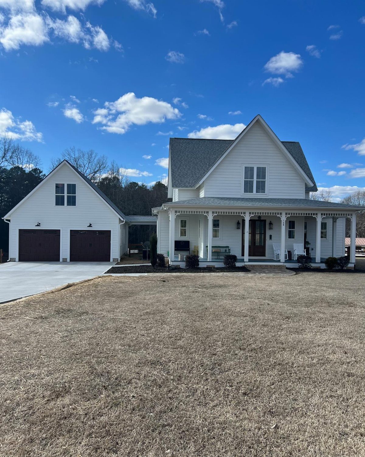 A white farmhouse with a large porch next to a detached garage under a blue sky.