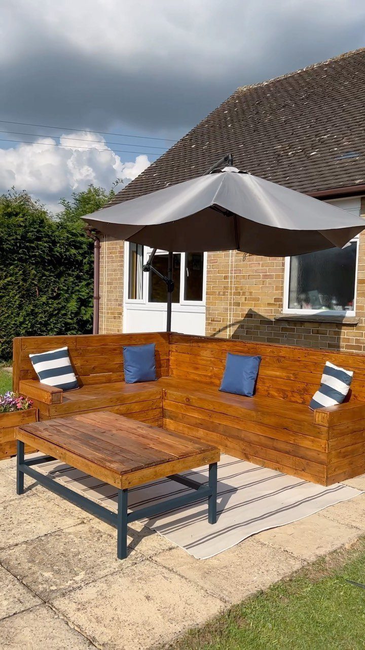 L-shaped wooden benches with blue cushions sit on a patio under a large grey umbrella.
