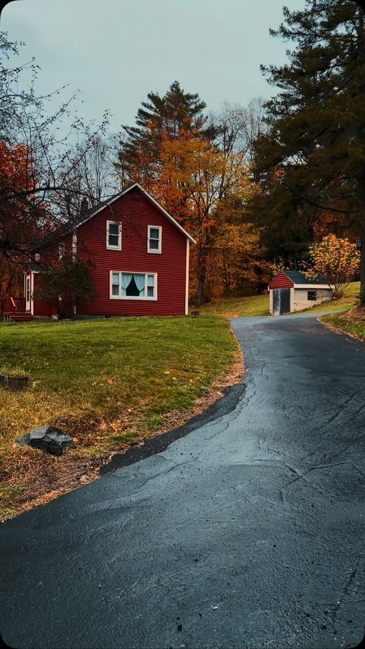 12 Trending House Colors for Exterior Looks in 2026 8 A red two-story house with white window trim surrounded by colorful autumn trees and a paved driveway.