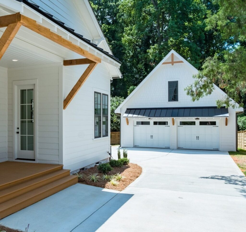 White farmhouse and detached garage with wooden beams and a concrete driveway under green trees.
