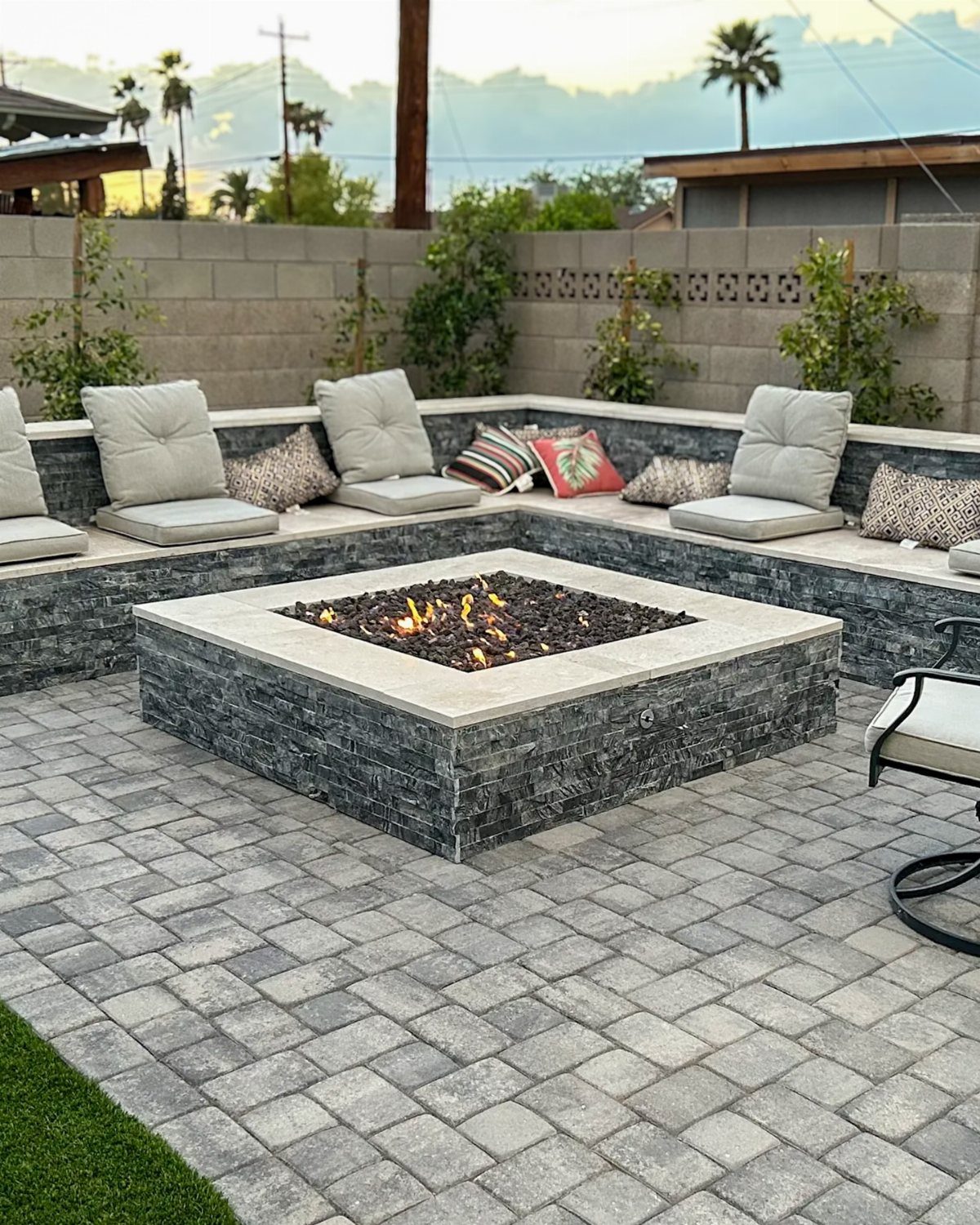 A square stone fire pit with built-in seating and cushions on a grey paver patio.