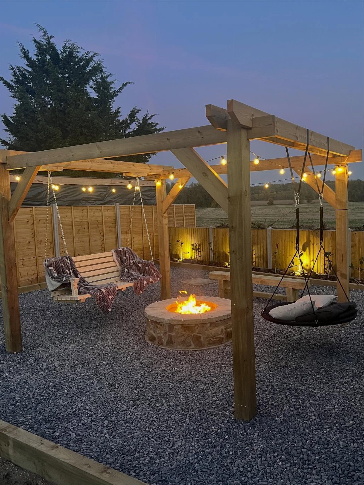 A stone fire pit burning brightly under a wooden pergola with hanging swings and string lights.