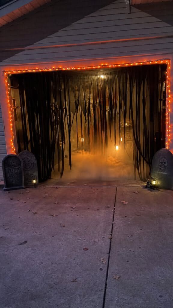 Halloween decorated garage entrance with black drapes, orange lights, gravestones, and low-lying fog.
