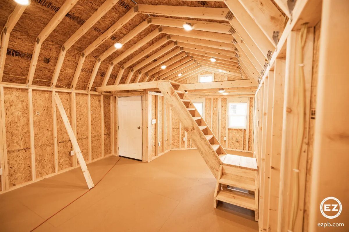 Unfinished wooden interior of a shed house featuring exposed framing, a loft, and a small staircase.