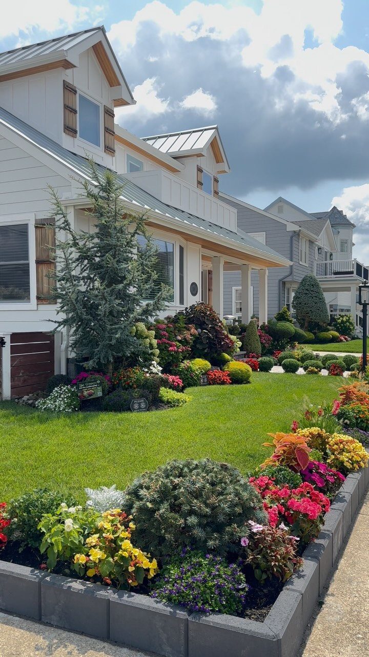Colorful flower beds with seasonal blooms and green shrubs line a manicured lawn in front of a house.