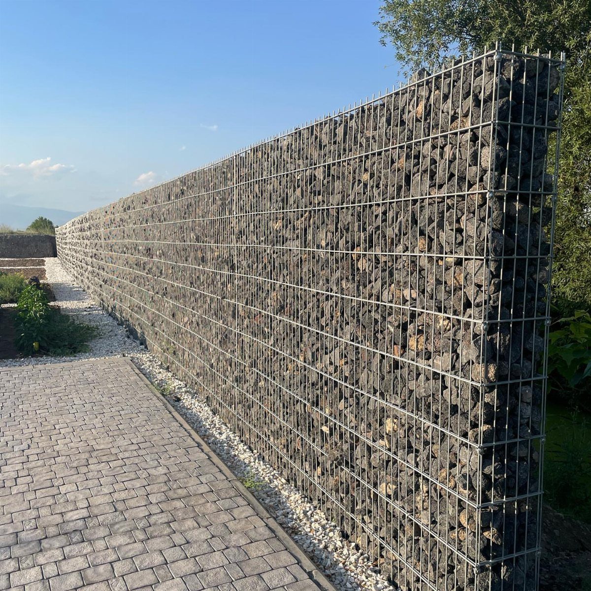 A long gabion wall filled with dark rocks alongside a grey stone paver walkway.