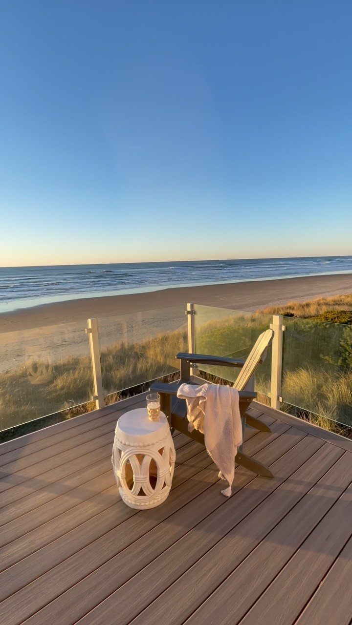 17 Beach House Deck Ideas for Oceanfront Living 14 A wooden deck with a glass railing overlooking a sandy beach and the ocean at sunset.