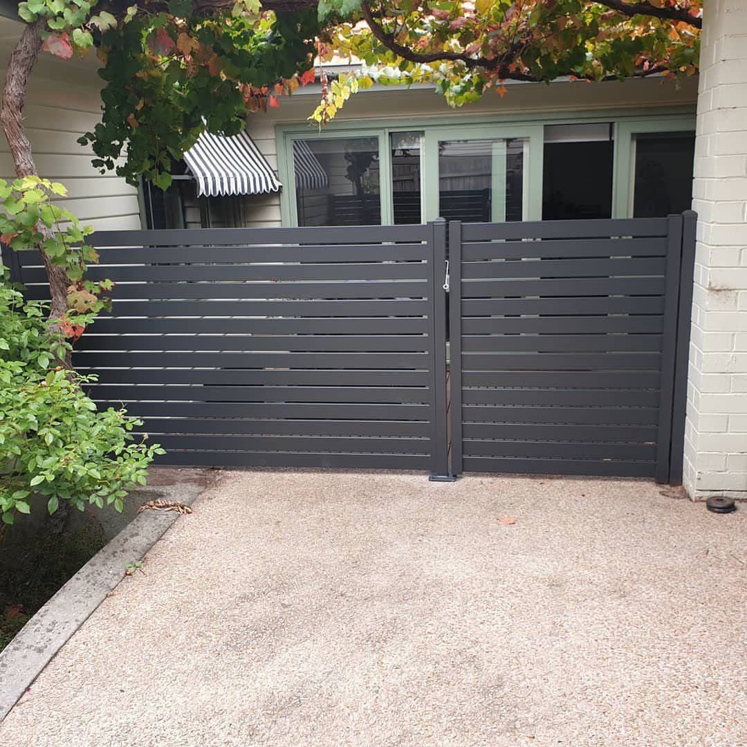 Dark grey horizontal slat fence and gate in front of a house with green plants.