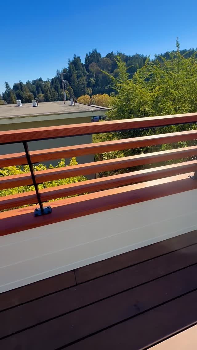14 Deck Balustrade Ideas for Every Style, from Glass to Stone 9 Horizontal reddish-brown wooden deck railing installed on a white base overlooking green trees.