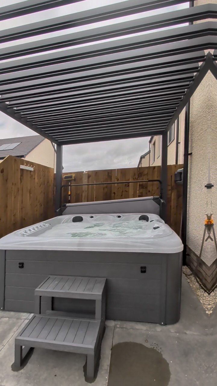 A grey hot tub sits under a dark metal pergola on a concrete patio.