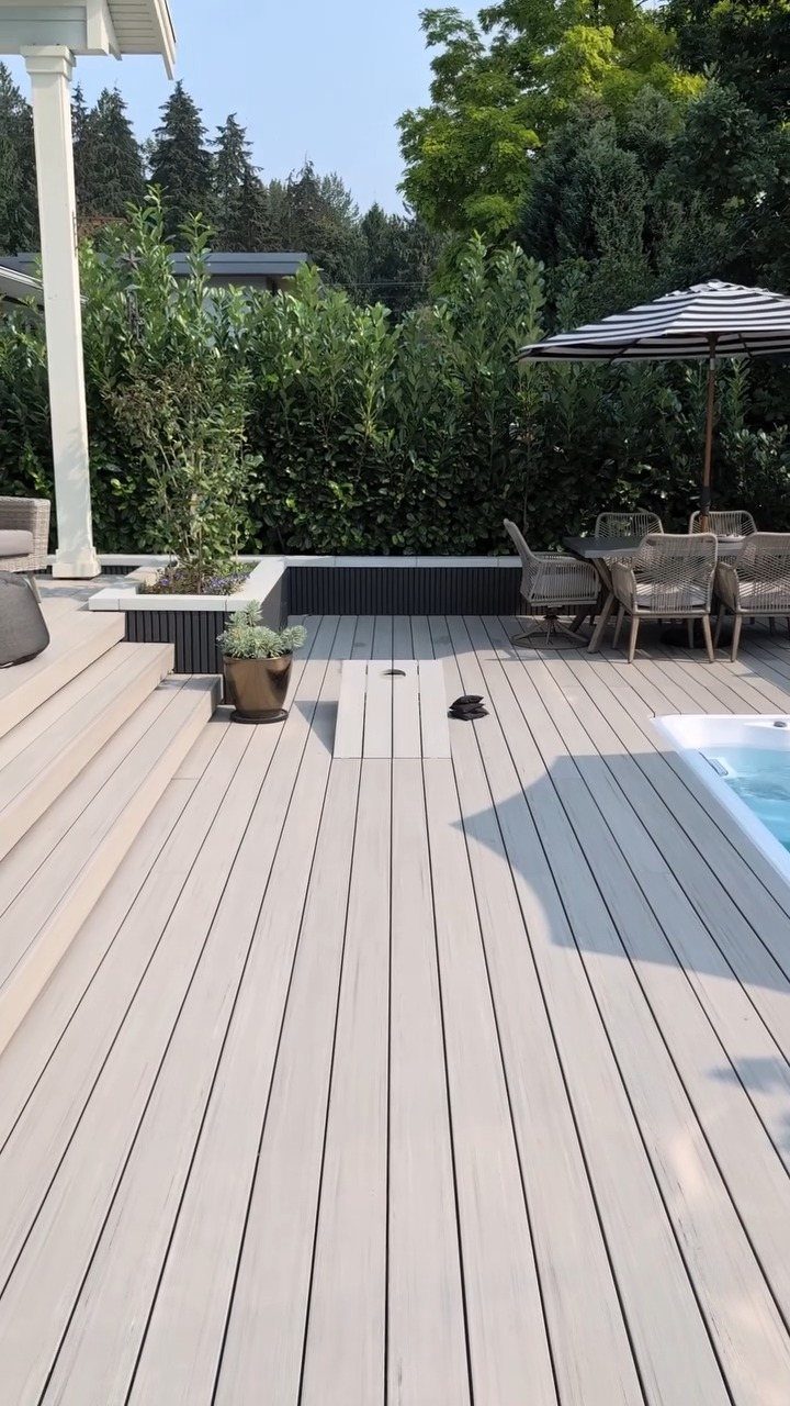 Light grey composite decking surrounding a swimming pool with outdoor patio furniture and green trees.