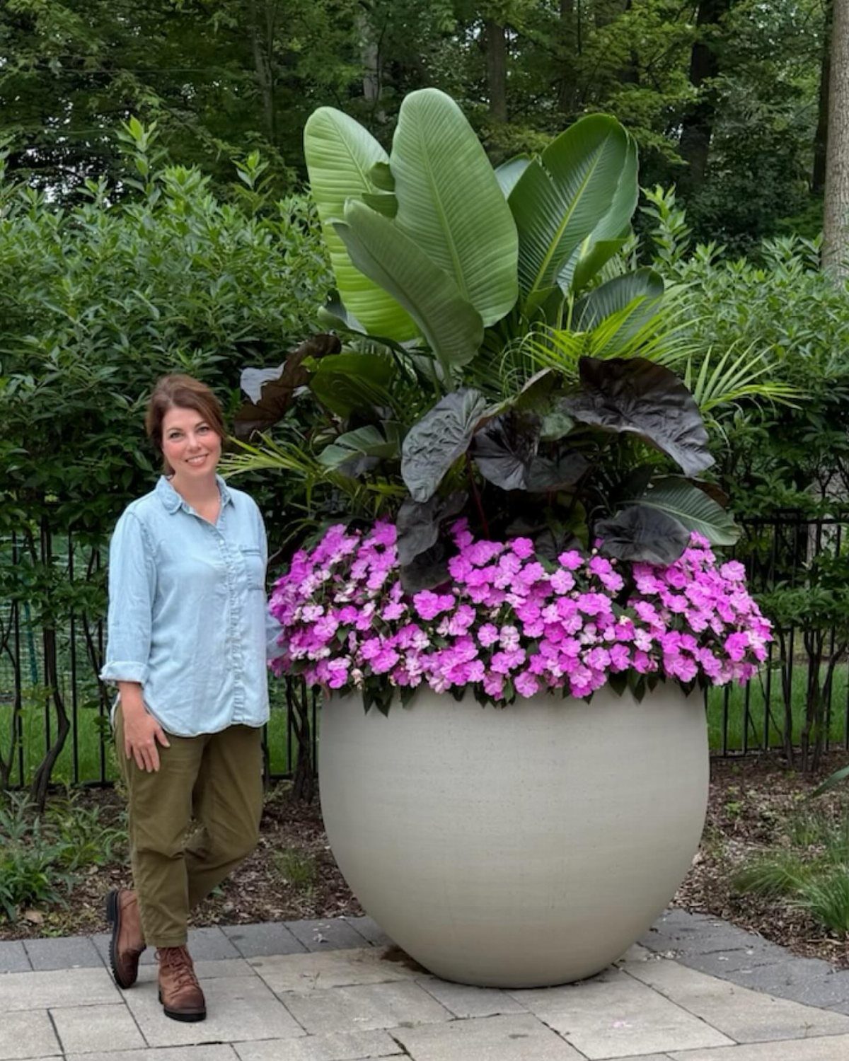 A woman stands beside a massive spherical planter filled with pink flowers and large tropical green leaves.