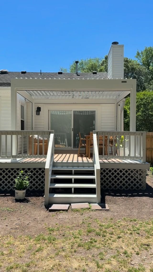 A light gray wooden deck with an attached pergola and outdoor dining set behind a white house.
