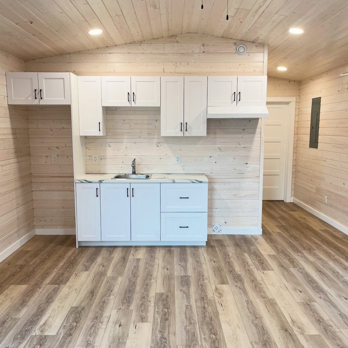 Interior of a small wooden shed house featuring white cabinets, a sink, and light wood paneling.