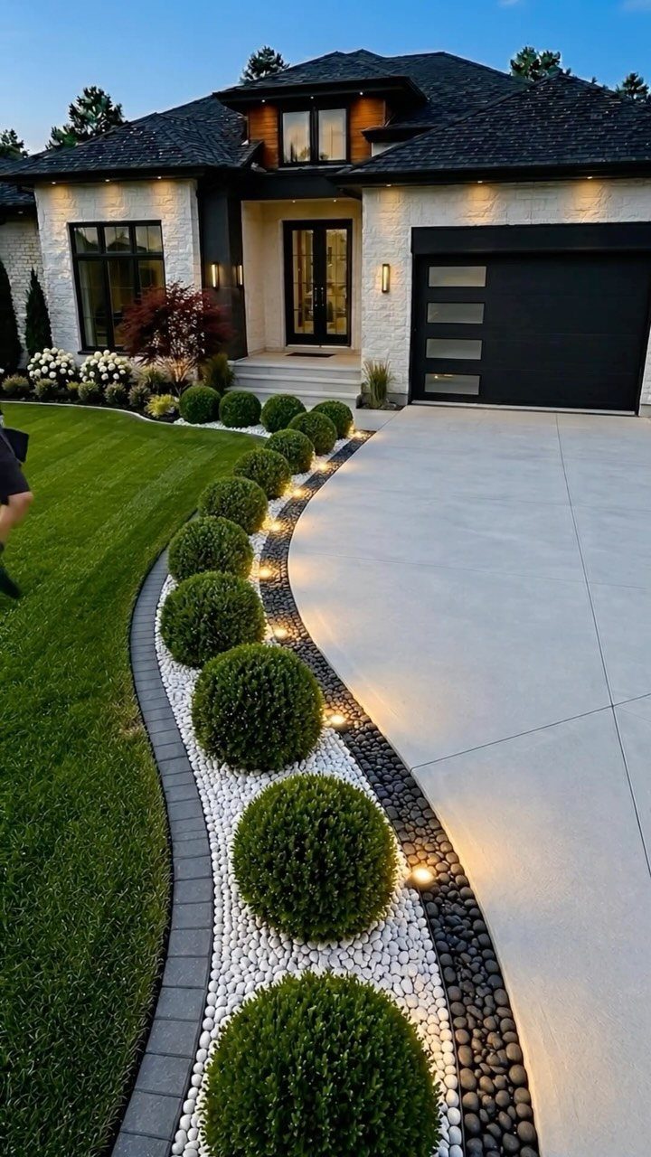Curved landscape bed with white river rocks, dark stones, round green shrubs, and small lights by a driveway.