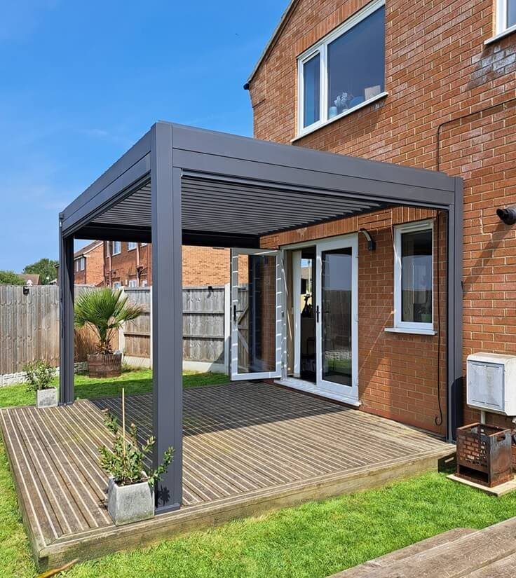 A dark grey louvered pergola attached to a brick house over a wooden deck.