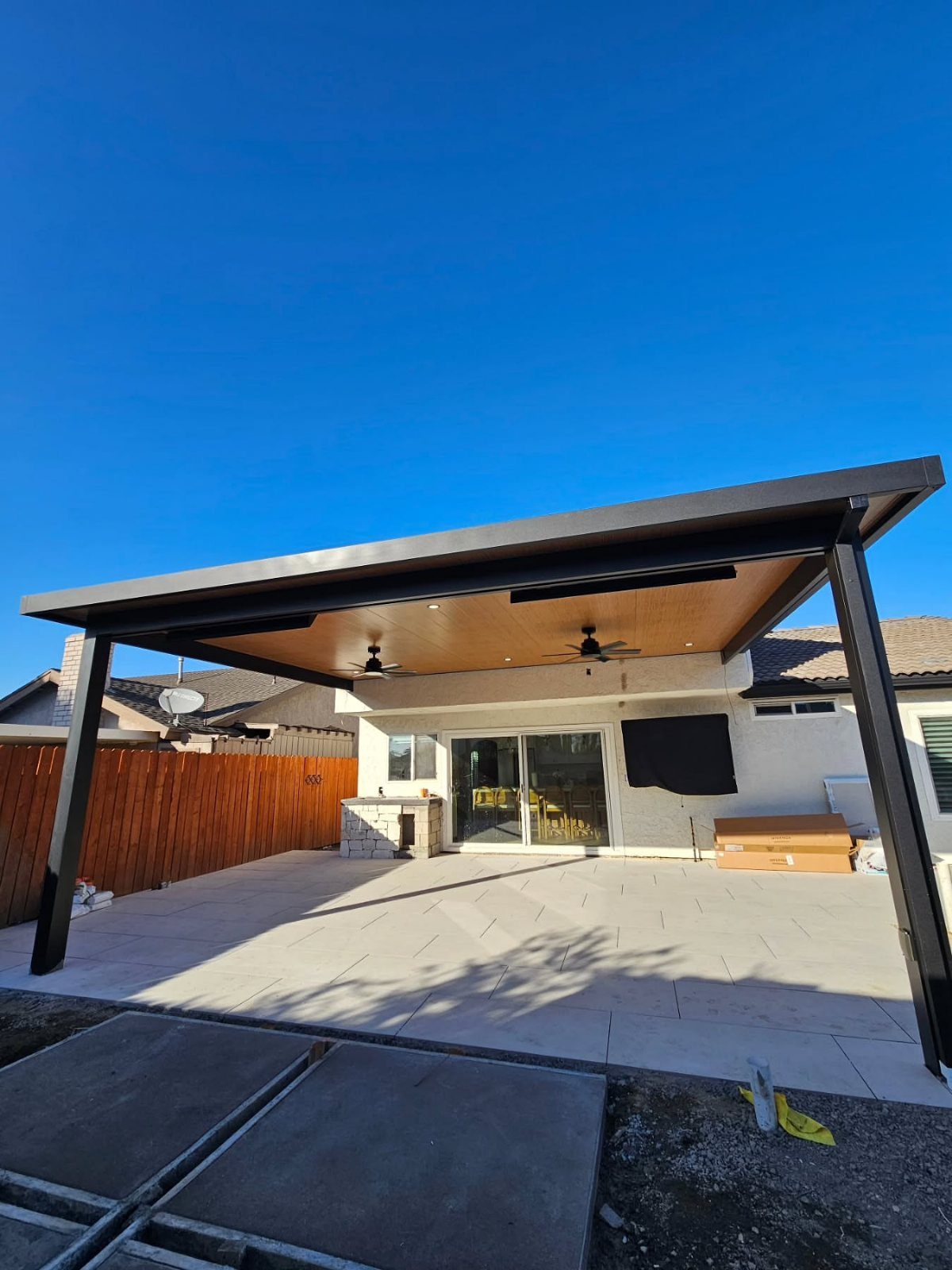 A contemporary dark metal patio cover with a wood-finished ceiling and ceiling fans over a stone patio.
