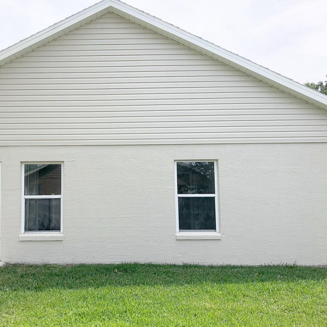 Light cream horizontal siding and stucco exterior wall with two white-trimmed windows and green grass.