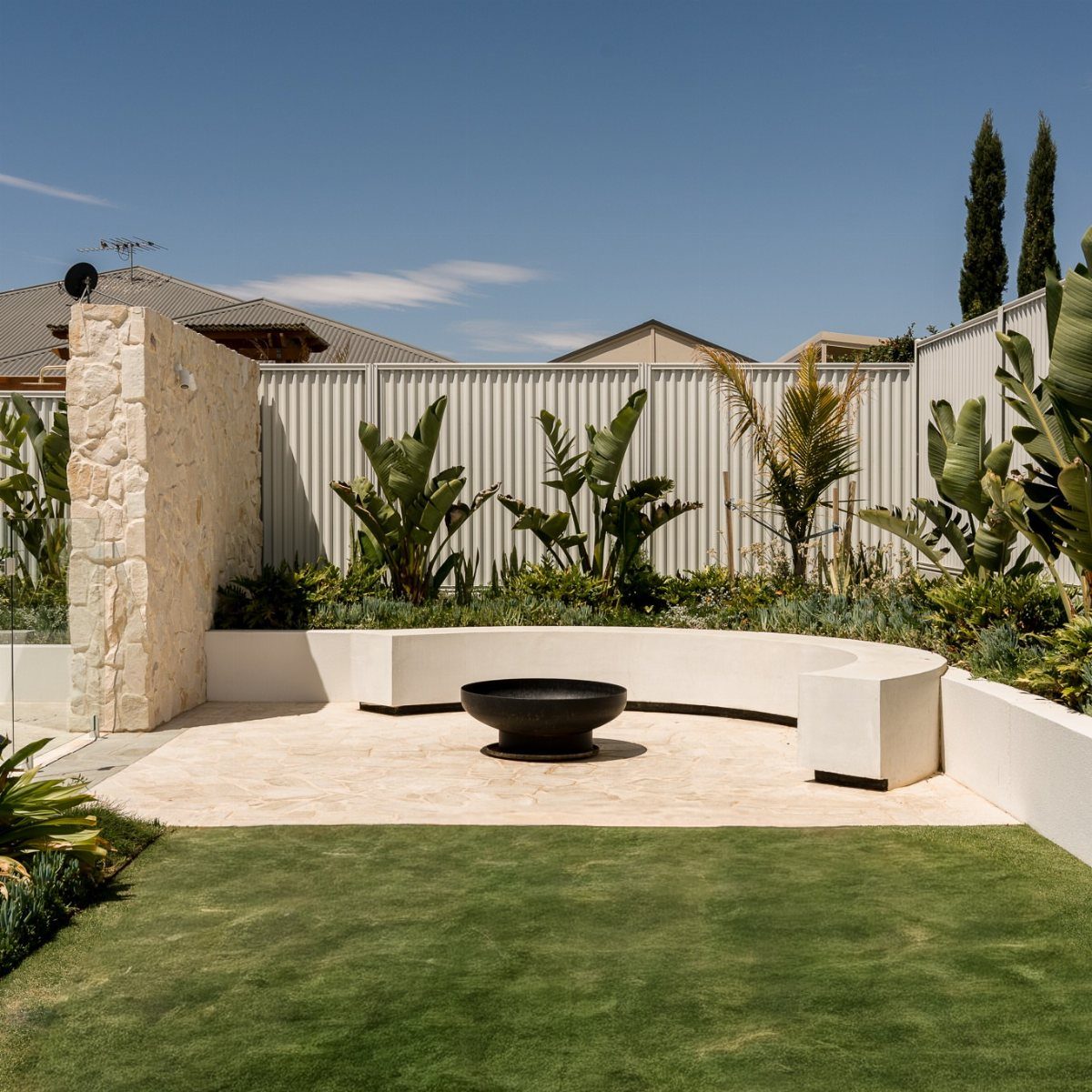 A black fire bowl on a stone patio surrounded by white curved benches and tropical plants.