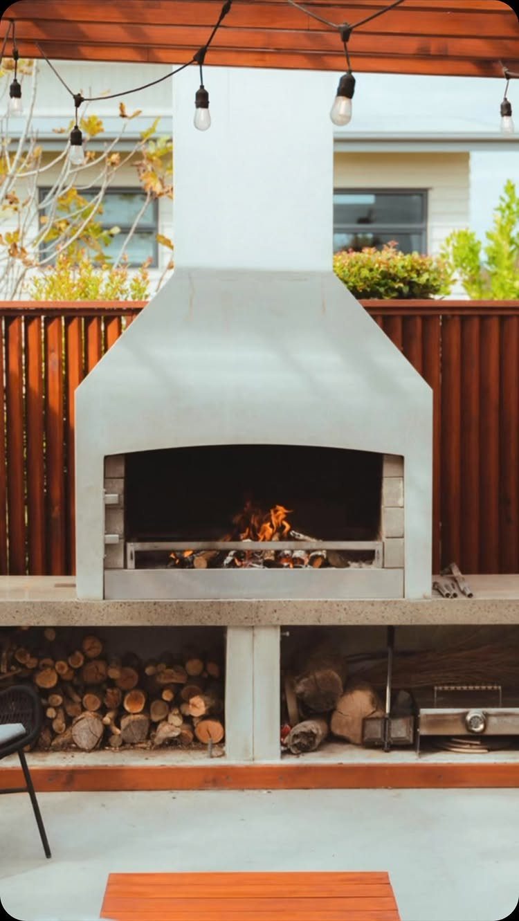 A modern concrete outdoor fireplace with a burning fire and stacked wood underneath.