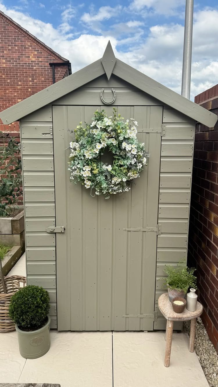 A sage green wooden garden shed with a white floral wreath on the door.