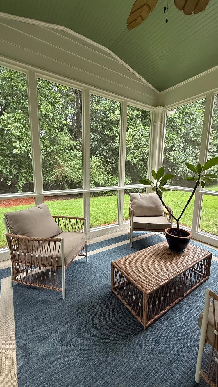 A furnished screened-in porch with wicker furniture, a blue rug, and views of green trees.