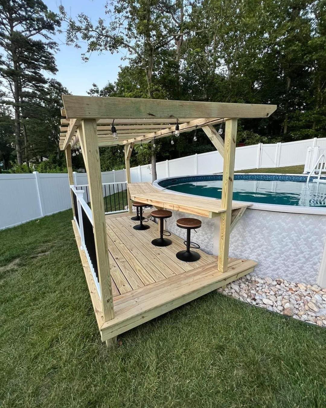 A wooden pergola bar structure with stools built on a deck next to a pool.