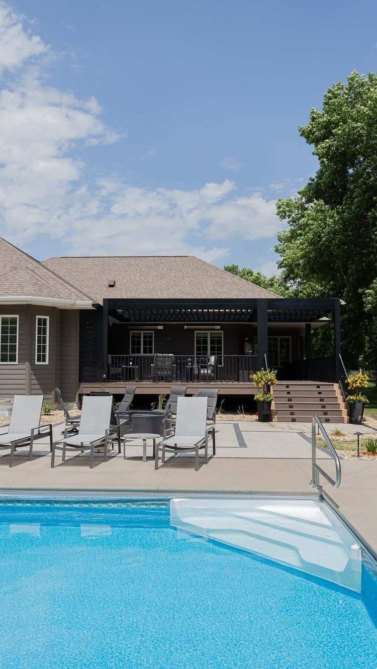 A wooden deck with a black pergola attached to a house next to a swimming pool.