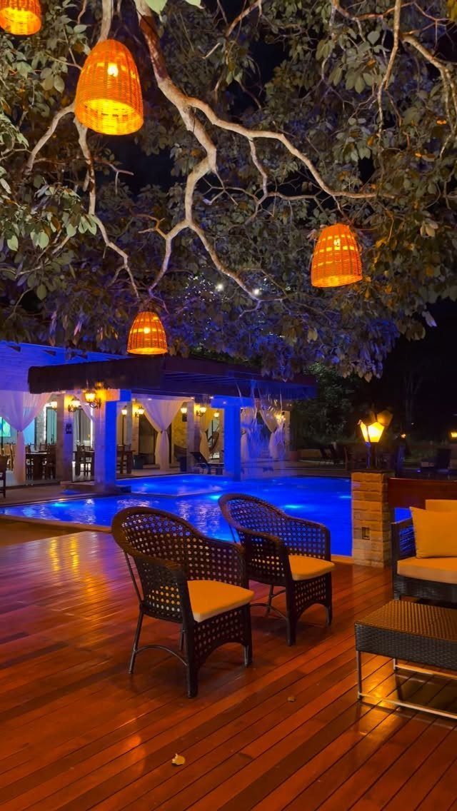 17 Beach House Deck Ideas for Oceanfront Living 13 Wicker chairs sit on a wooden deck near a glowing blue swimming pool at night.