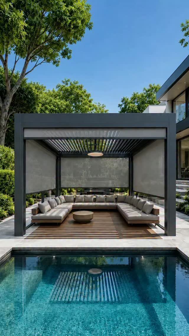 A modern dark grey pergola with a grey sectional sofa sits beside a blue swimming pool.