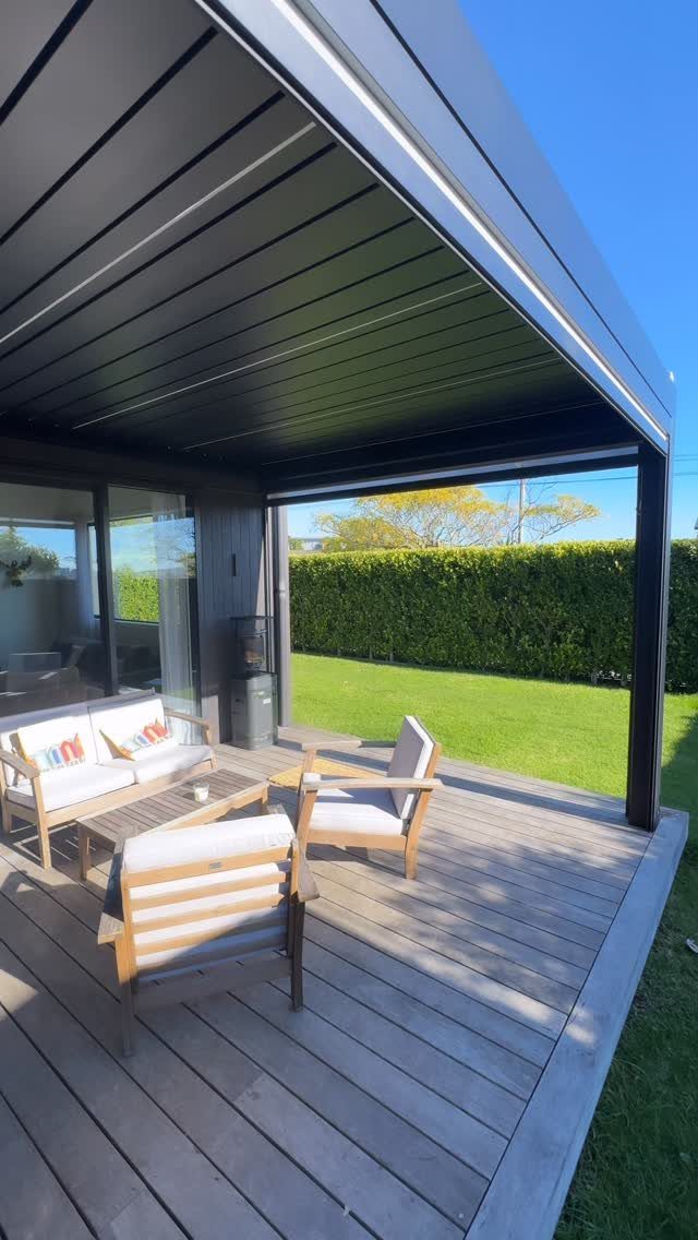 A modern black pergola with a retractable roof covers a wooden deck with outdoor lounge furniture.