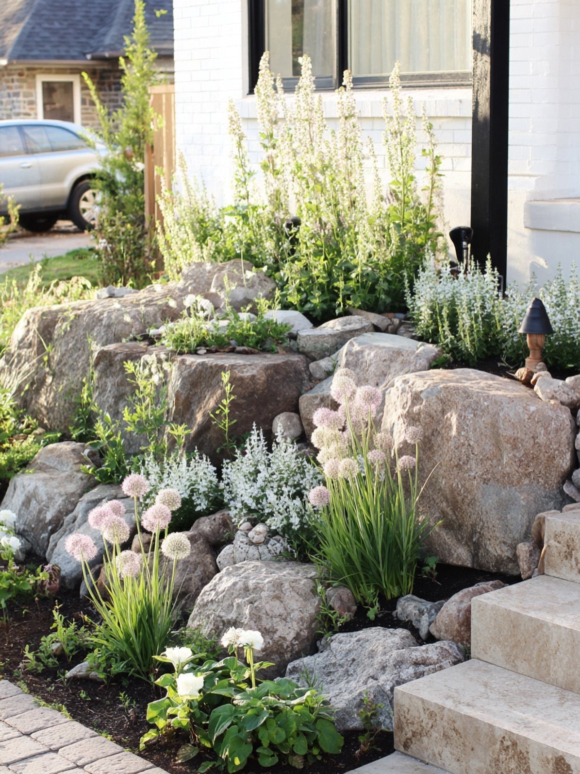 Large landscape boulders and various green plants with white flowers in a front yard garden bed.