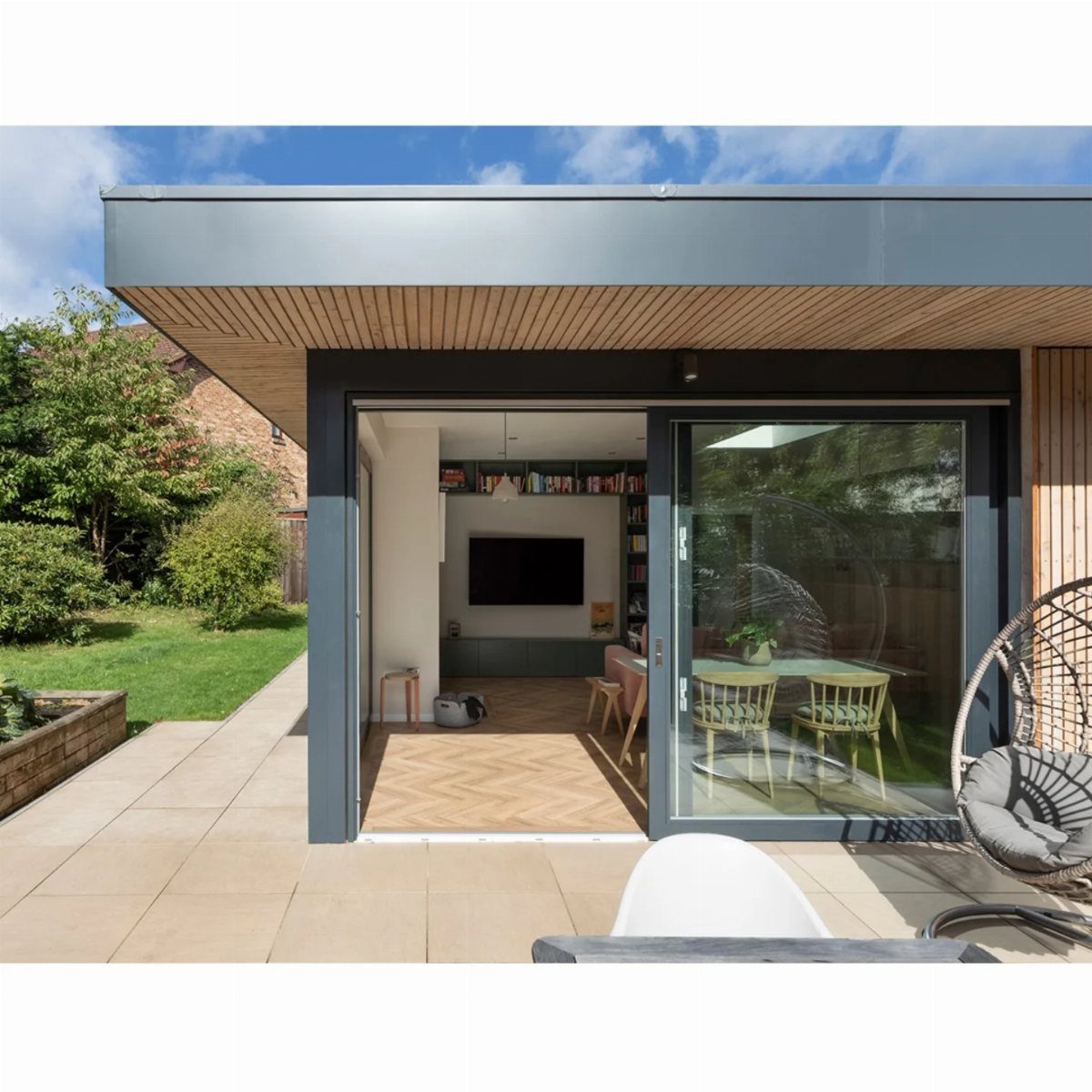 Modern house extension featuring a large flat roof overhang with light wood slats above a stone patio.