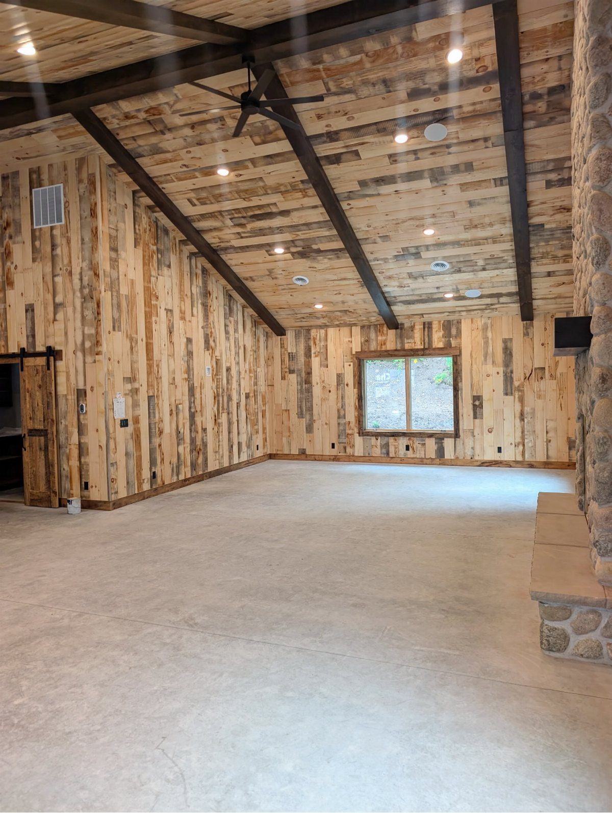 Empty room with rustic wood paneled walls, vaulted ceiling, dark beams, and concrete flooring.