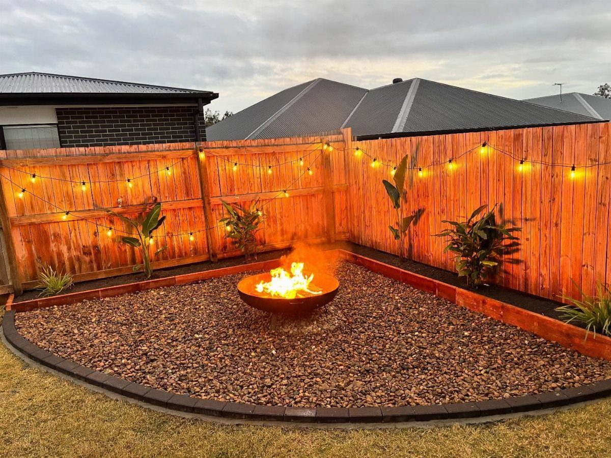 A burning fire bowl sits in a gravel backyard area surrounded by a wooden fence and string lights.