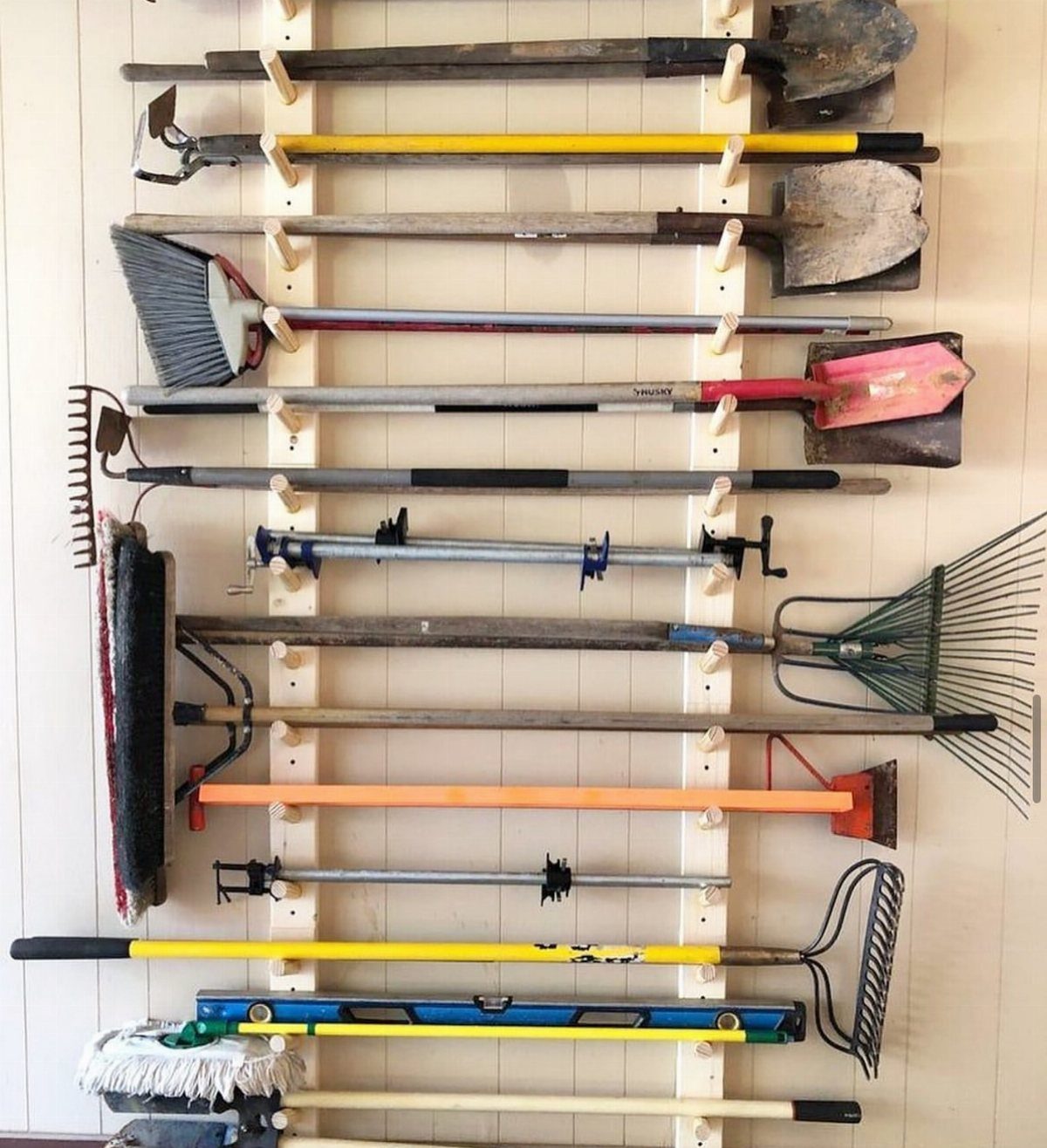 Long-handled yard tools organized on white wall-mounted racks against a light paneled wall.
