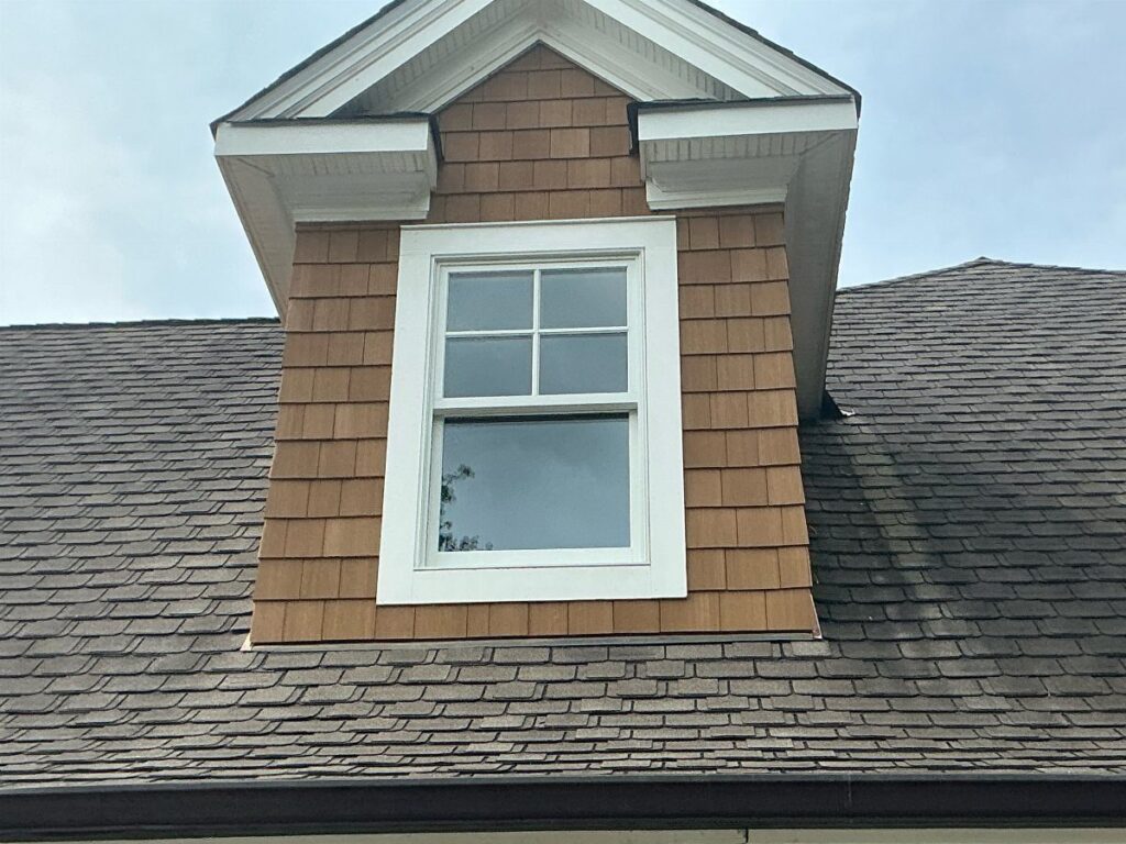 Brown cedar shake siding on a house dormer with a white window and grey roof shingles.