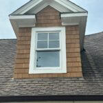 Brown cedar shake siding on a house dormer with a white window and grey roof shingles.