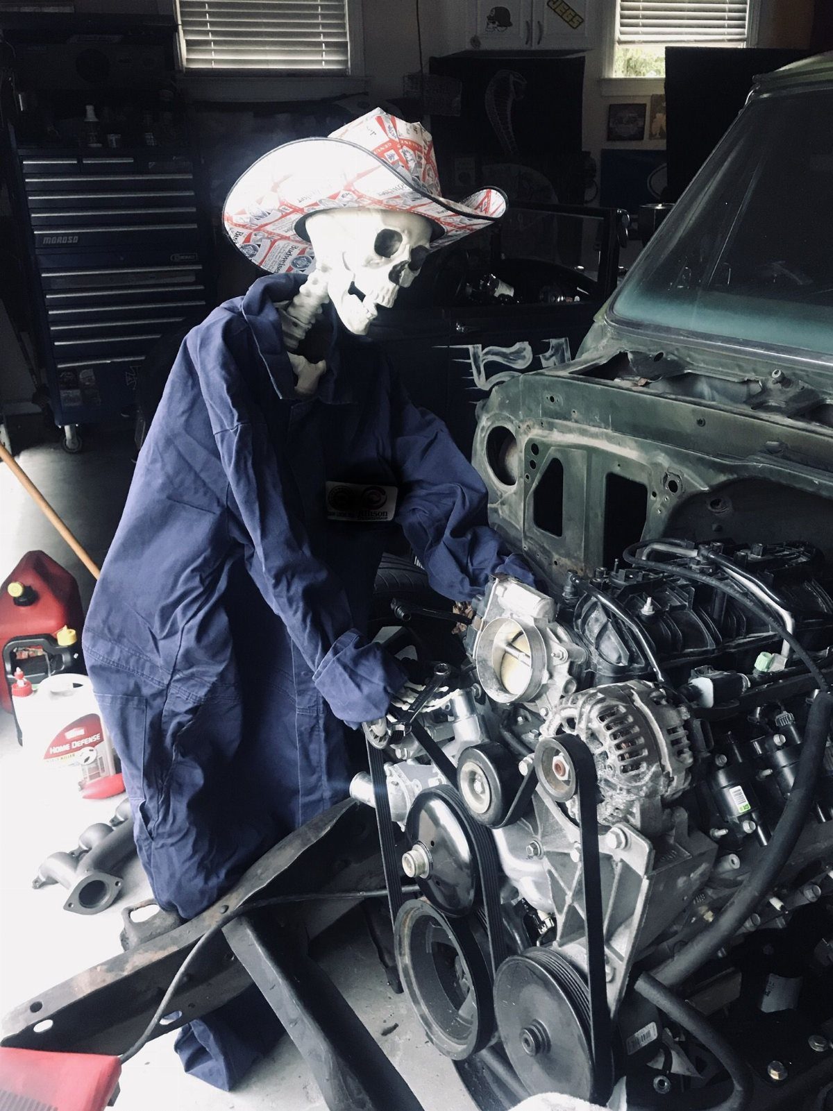 Skeleton wearing a blue jumpsuit and cowboy hat working on a car engine in a garage.