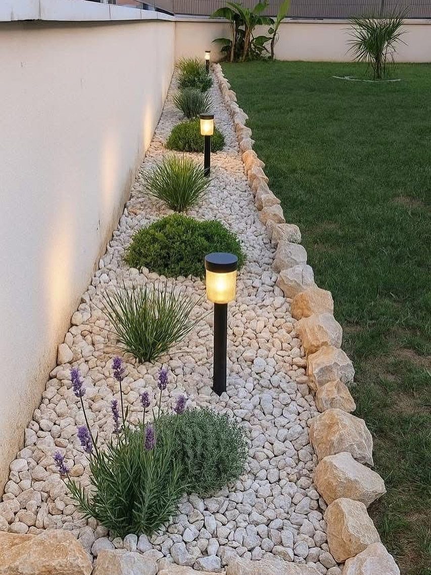 A narrow garden bed with white pebbles, green shrubs, and solar lights edged by natural stones.